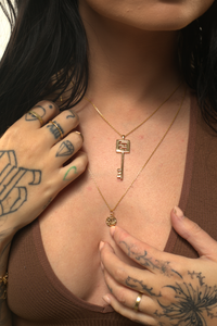 A person wearing a brown top and showcasing a gold-colored necklace with a key-shaped pendant and a unique logo tag, along with other gold jewelry.