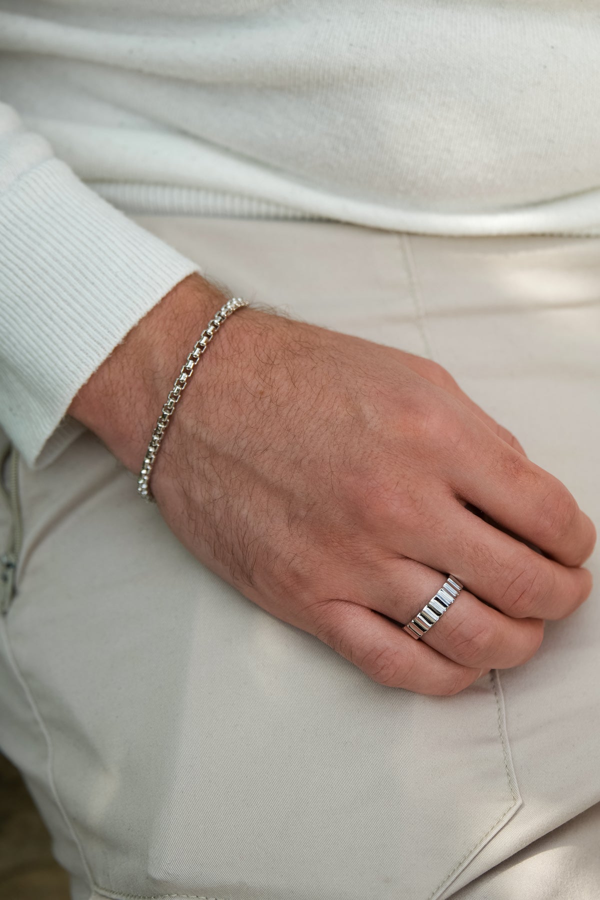 Hand wearing a silver ring and box chain bracelet with a white sweater and beige pants